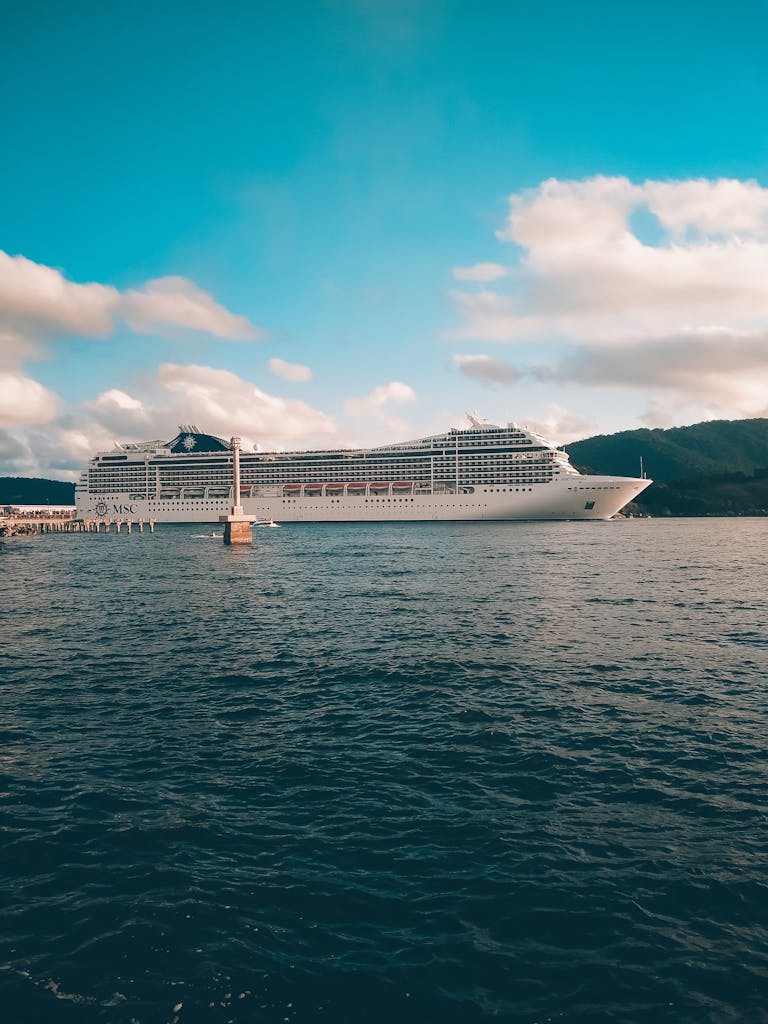 A grand cruise ship sails under a bright blue sky with fluffy clouds over a calm ocean.