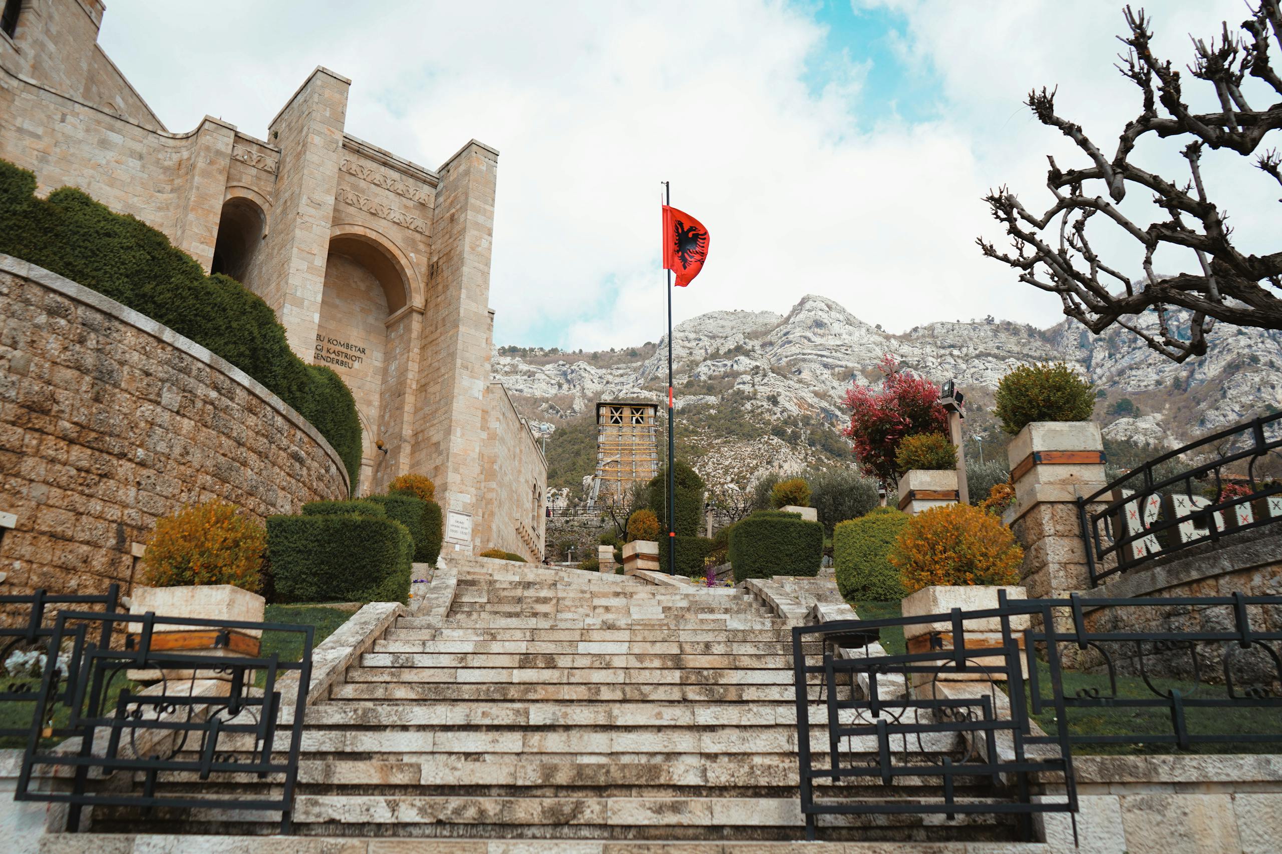 Stone steps leading to a castle with the Albanian flag, set against mountainous terrain.